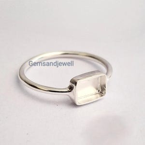 May include: A silver-colored ring featuring a rectangular bezel setting designed for a gemstone. The metal band is thin and smooth, and the text Gemsandjewell is visible on the surface near the setting. The ring has a polished finish.