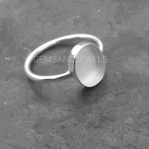 Può includere: Un anello d'argento con una grande pietra ovale di colore chiaro. La fascia dell'anello si curva per incontrare l'incastonatura della pietra. Il testo "GEMSANDJEWELS" è visibile sull'anello.
