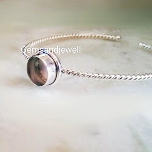 May include: A silver bangle bracelet with a round bezel setting for a gemstone. The bracelet has a twisted rope design.