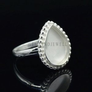 May include: A silver ring with a pear-shaped, white gemstone. The gemstone is surrounded by a beaded border. The ring is set against a black background, with a reflection of the ring visible below. The text "GEMANDJEWELL" is visible.
