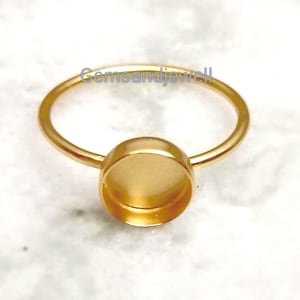 May include: A gold-toned metal ring featuring a circular bezel setting designed for a cabochon or stone. The ring has a thin, smooth band and a flat, recessed cup at the center. The text Gemsandjewell is visible in blue letters above the ring.