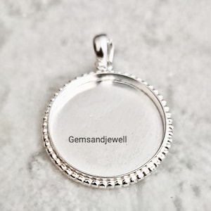 May include: A circular silver pendant tray featuring a beaded edge and a smooth, flat centre surface for customisation. The pendant includes a small bail at the top for attachment. The text Gemsandjewell is printed across the centre of the piece.