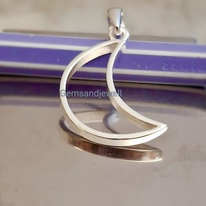 May include: A silver-colored crescent moon pendant. The pendant is a minimalist design, with a smooth, polished surface. The moon shape is open, with a slight curve. The pendant has a small loop at the top for a chain. The background is a reflective surface.