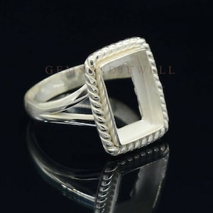 May include: A silver ring with a rectangular setting, featuring a rope-like border. The ring's band is smooth and simple, with a split design near the setting. The ring is displayed against a dark background, highlighting its reflective surface.