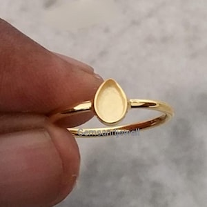 May include: A gold ring featuring a teardrop-shaped setting. The ring has a slender band and a simple, elegant design. The ring is held by a hand.