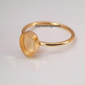 Peut inclure: Une bague dorée avec un sertissage de lunette ovale. L'anneau de la bague est fin et lisse, et la lunette est conçue pour accueillir une pierre précieuse ou un autre élément décoratif. Le texte "gemsandjewell" est imprimé sur l'anneau.