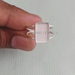 Può includere: Un anello d'argento con una gemma rettangolare rosa chiaro. L'anello ha un design a doppia fascia. Il testo "GEMS AND JEWELL" è visibile sull'anello.
