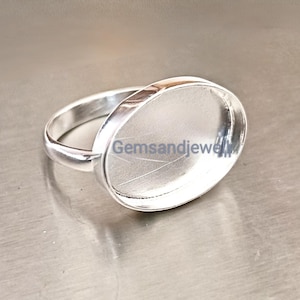 May include: A silver ring with an oval bezel setting. The ring is made of a shiny, metallic material and is set against a neutral background. The text "Gemsandjewel" is visible on the ring.