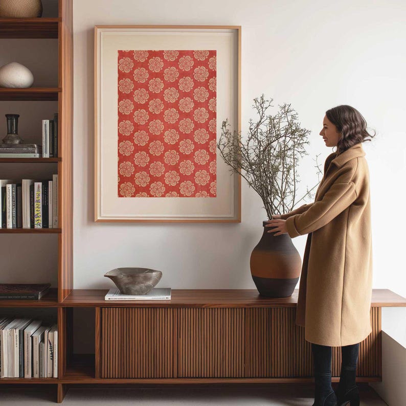 Vintage fabric wall art print with a red floral block print stencil pattern, framed and displayed in a home office beside a bookcase and a woman arranging branches in a clay vase.