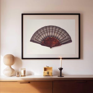 May include: A framed print of a vintage fan with a dark lace design and a wooden handle. The fan is displayed on a white background. A wooden cabinet with decorative items, including a candle and a small wooden box, is in the foreground.