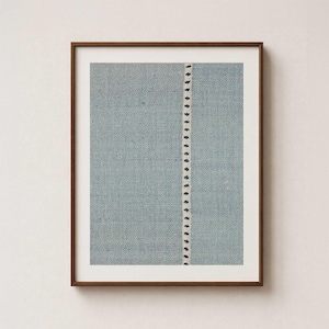 May include: A framed wall art print featuring a textured light blue fabric background with a repeating geometric pattern and a single vertical off-white stripe down the center containing small, dark blue, evenly spaced floral-like motifs.