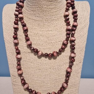 Puede incluir: Un collar largo con cuentas redondas de varios tamaños y un patrón moteado en tonos burdeos intenso, marrón oscuro y blanco crema. Las cuentas están ensartadas en un bucle continuo, creando un efecto de capas con un acabado liso y pulido.