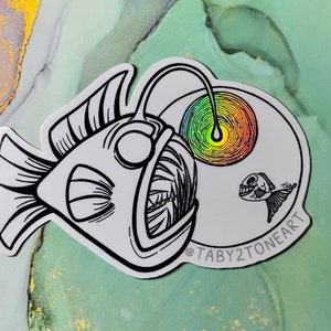 Puede incluir: Un dibujo en blanco y negro de un pez pescador con una boca grande y un señuelo. El pez pescador está nadando hacia un pez más pequeño. El fondo es gris claro con un remolino de arcoíris.
