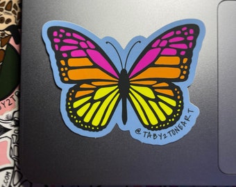 May include: A sticker of a colorful butterfly with yellow, orange, pink, and black wings. The butterfly is on a light blue background with the text "@tabby2toneart" at the bottom.