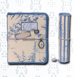 Op de afbeelding: Een blauw en crèmekleurig notitieboek- en pennenhouder set. Het notitieboek heeft een bloemenontwerp met tekst en een hanger. De pennenhouder heeft blauwe en crèmekleurige strepen. De set wordt bij elkaar gehouden door een grijze elastische band.