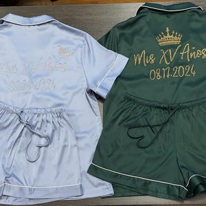 May include: Two pairs of satin pajamas with short sleeves and shorts. One pair is light blue with the text "Mis XV Años 08.17.2024" embroidered on the back. The other pair is dark green with a gold crown and the text "Mis XV Años 08.17.2024" embroidered on the back.