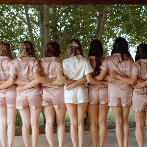 May include: A group of eight people wearing matching pink satin pajamas with embroidered text on the back. The text reads "Bride", "Bridesmaid", "Maid of Honor", and "Bridesquad".