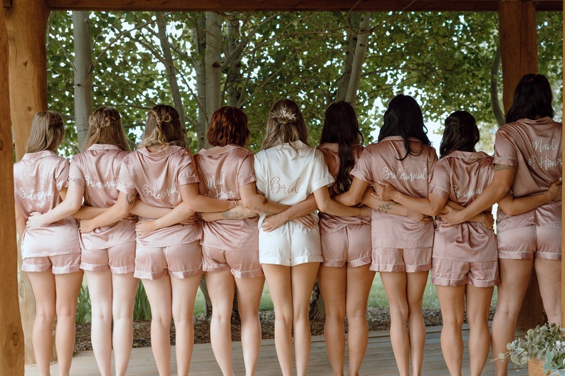 May include: A group of eight people wearing matching pink satin pajamas with embroidered text on the back. The text reads "Bride", "Bridesmaid", "Maid of Honor", and "Bridesquad".