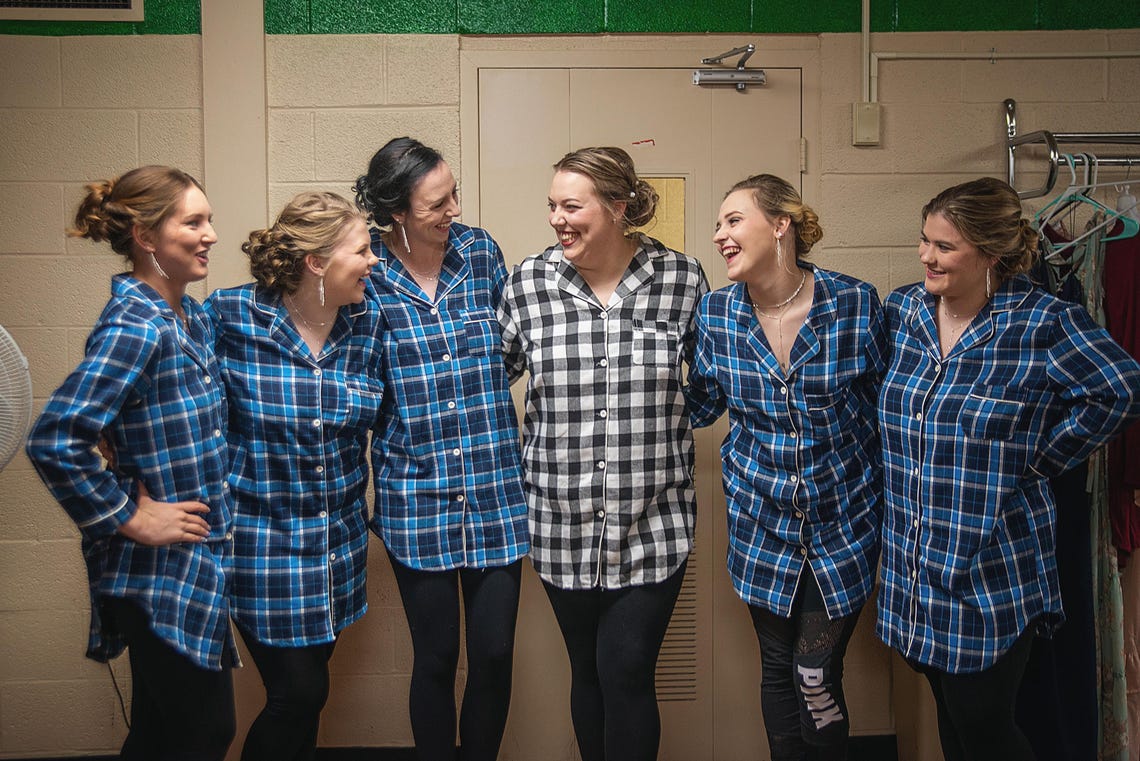 May include: Six bridesmaids pose in matching blue and white plaid pajama shirts.  The shirts have a button-down front and long sleeves. Two bridesmaids wear black and white plaid shirts. One bridesmaid's leggings say 'PINK' on the leg. The group is standing in a room with a green wall and a door.