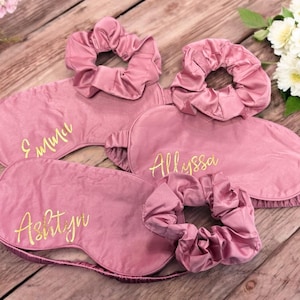 May include: Pink satin sleep masks and matching scrunchies. Each mask is embroidered with a name in gold script: Emma, Allyssa, and Ashtyn. The items are arranged on a wooden surface with flowers.