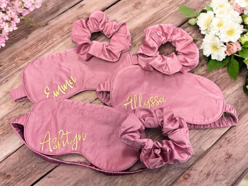 May include: Pink satin sleep masks and matching scrunchies. Each mask is embroidered with a name in gold script: Emma, Allyssa, and Ashtyn. The items are arranged on a wooden surface with flowers.