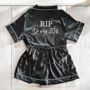 May include: Black satin short-sleeved pajama top and shorts set with white piping. The top has the text "RIP to my 20's" printed on the back in rose gold.