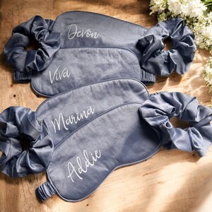 May include: Four grey satin sleep masks with matching scrunchies. Each mask is embroidered with a name in white script: Devon, Viva, Marina, and Addie. The items are arranged on a wooden surface, with white flowers in the background.