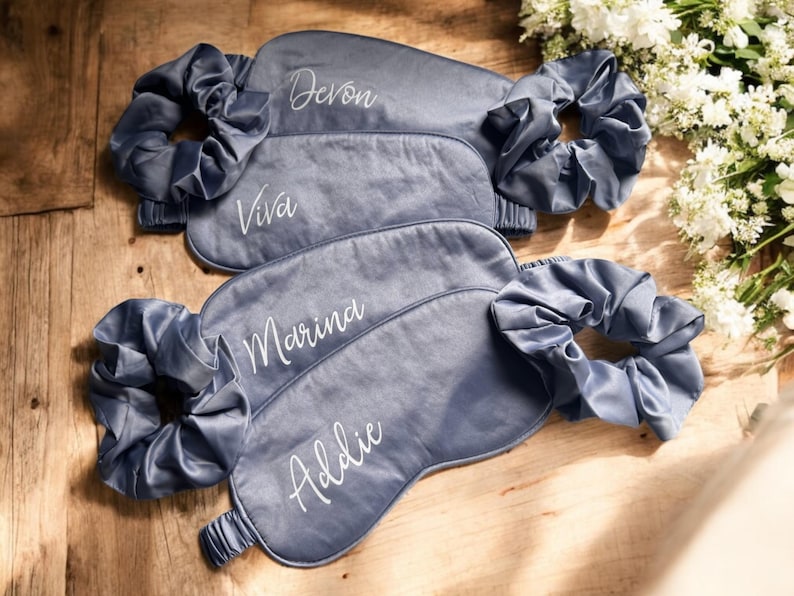 May include: Four grey satin sleep masks with matching scrunchies. Each mask is embroidered with a name in white script: Devon, Viva, Marina, and Addie. The items are arranged on a wooden surface, with white flowers in the background.