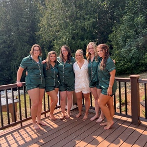May include: Six women wearing green satin pajamas with white trim and gold lettering. They are standing on a wooden deck with a railing in the background.