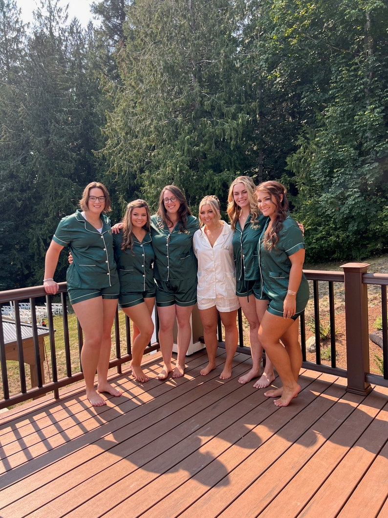 May include: Six women wearing green satin pajamas with white trim and gold lettering. They are standing on a wooden deck with a railing in the background.