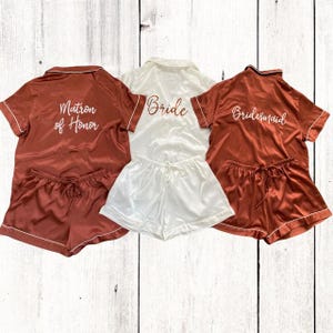 May include: Three sets of matching pajamas with short sleeves and shorts. The pajamas are in a rust color with white piping. The tops have the words "Bride", "Matron of Honor", and "Bridesmaid" embroidered on the back in white script.