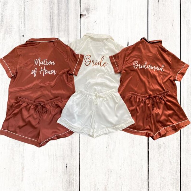 May include: Three sets of matching pajamas with short sleeves and shorts. The pajamas are in a rust color with white piping. The tops have the words "Bride", "Matron of Honor", and "Bridesmaid" embroidered on the back in white script.