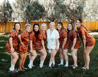 May include: A group of eight people wearing matching rust-colored satin pajamas with white trim. The people are standing in a line, smiling at the camera. They are all wearing white socks and some are wearing sandals.