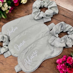 May include: Three grey satin sleep masks and matching scrunchies are arranged on a wooden surface. Each mask is personalised with a name in white script: Julia, Dara, and Aldeze. The masks and scrunchies are a light grey colour.