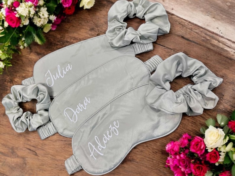 May include: Three grey satin sleep masks and matching scrunchies are arranged on a wooden surface. Each mask is personalised with a name in white script: Julia, Dara, and Aldeze. The masks and scrunchies are a light grey colour.
