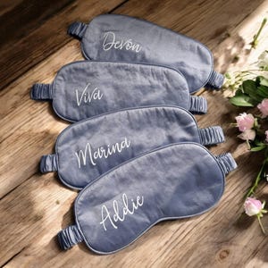 May include: Four grey sleep masks, each embroidered with a name in white script: Devon, Viva, Marina, and Addie. The masks are on a wooden surface, with a bouquet of pink and white flowers in the background.