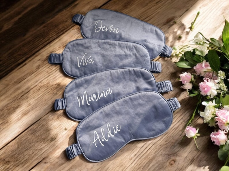 May include: Four grey sleep masks, each embroidered with a name in white script: Devon, Viva, Marina, and Addie. The masks are on a wooden surface, with a bouquet of pink and white flowers in the background.