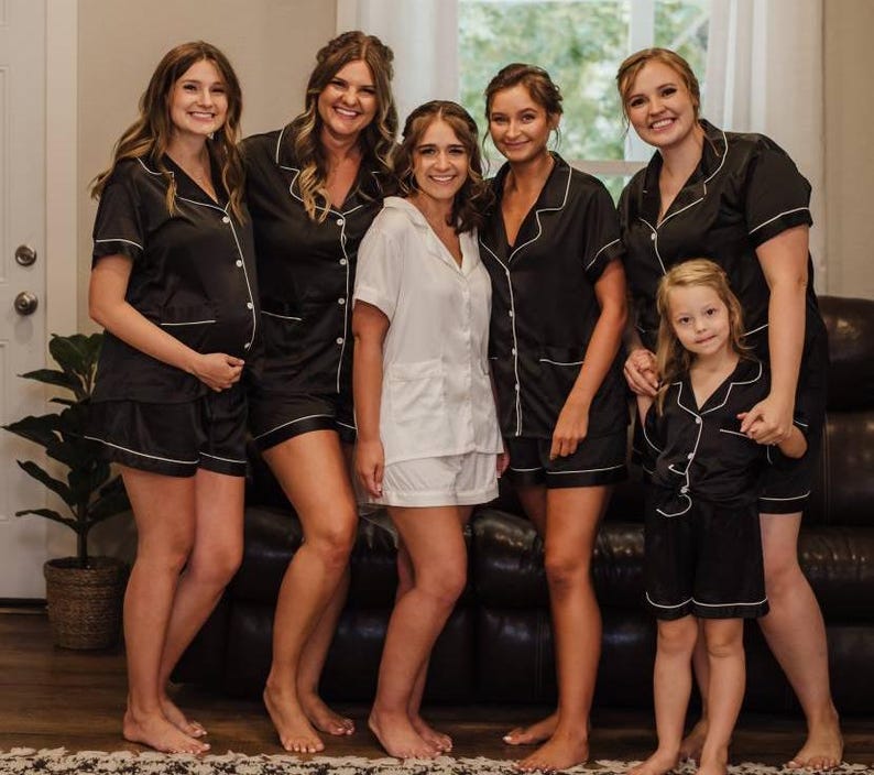 May include: Six people wearing black and white pajamas pose for a photo. The women are wearing black satin pajamas with white piping. The girl is wearing a black satin pajama set with white piping. The women are smiling and looking at the camera.