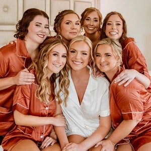 May include: A group of seven women wearing matching rust-colored satin pajamas with white piping. The women are smiling and posing for a photo. The bride is in the center wearing a white satin pajama set.