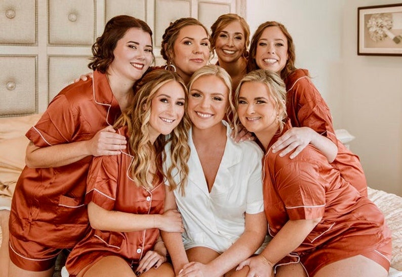 May include: A group of seven women wearing matching rust-colored satin pajamas with white piping. The women are smiling and posing for a photo. The bride is in the center wearing a white satin pajama set.