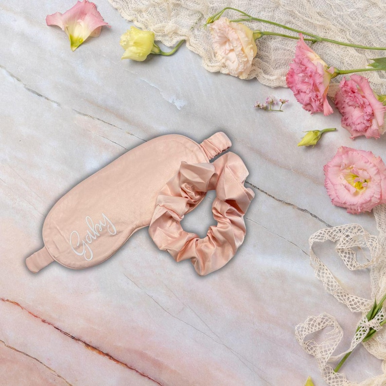 May include: A pale pink sleep mask and scrunchie set. The sleep mask is embroidered with the name "Gaby" in white script. The set is presented on a marble surface, decorated with pink flowers and lace.