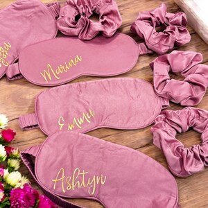 May include: Mauve sleep masks and scrunchies, each with a gold script name. The masks are embroidered with the names "Alyssa", "Marina", "Emma", and "Ashtyn". The items are arranged on a wooden surface, with a floral bouquet. A set of accessories for relaxation.