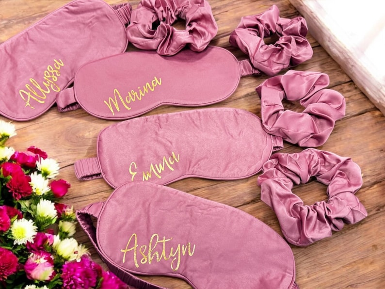 May include: Mauve sleep masks and scrunchies, each with a gold script name. The masks are embroidered with the names "Alyssa", "Marina", "Emma", and "Ashtyn". The items are arranged on a wooden surface, with a floral bouquet. A set of accessories for relaxation.
