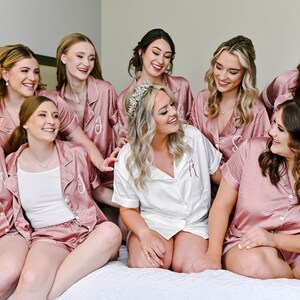 May include: A group of women in matching pink satin pajamas, with one woman in the center wearing a white satin pajama top and a tiara. They are all smiling and looking at each other.