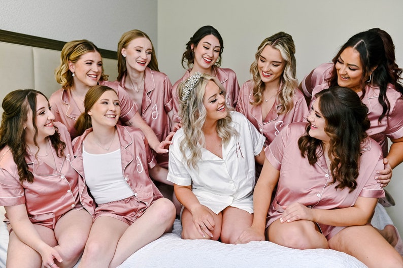 May include: A group of women in matching pink satin pajamas, with one woman in the center wearing a white satin pajama top and a tiara. They are all smiling and looking at each other.