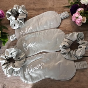 May include: A set of three gray satin sleep masks and matching scrunchies, each personalized with a name in white script: Julia, Dara, and Adaeze. The items are arranged on a wooden surface, with floral arrangements in the background.