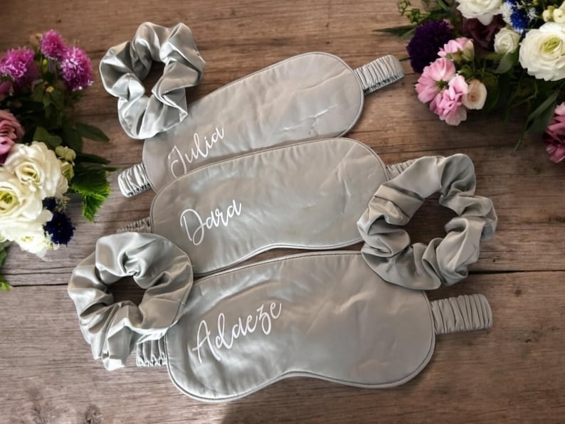 May include: Three grey satin sleep masks and matching scrunchies, each personalised with a name in white script: Julia, Dara, and Adaeze. The items are arranged on a wooden surface, with floral arrangements in the background.