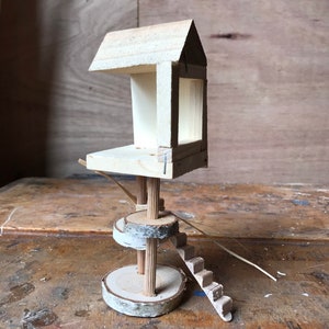 May include: A miniature wooden treehouse with a roof, a platform, and a ladder. The treehouse is built on two wooden discs with a birch bark finish.