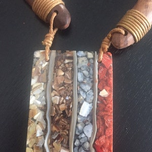 May include: A rectangular pendant necklace with a wood and rope design. The pendant is made of different coloured stones and is set in a clear resin.