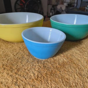 May include: Three vintage mixing bowls in yellow, teal, and blue, each with a white interior. The bowls are nested on a textured, golden-brown surface. The bowls are a classic kitchenware item.